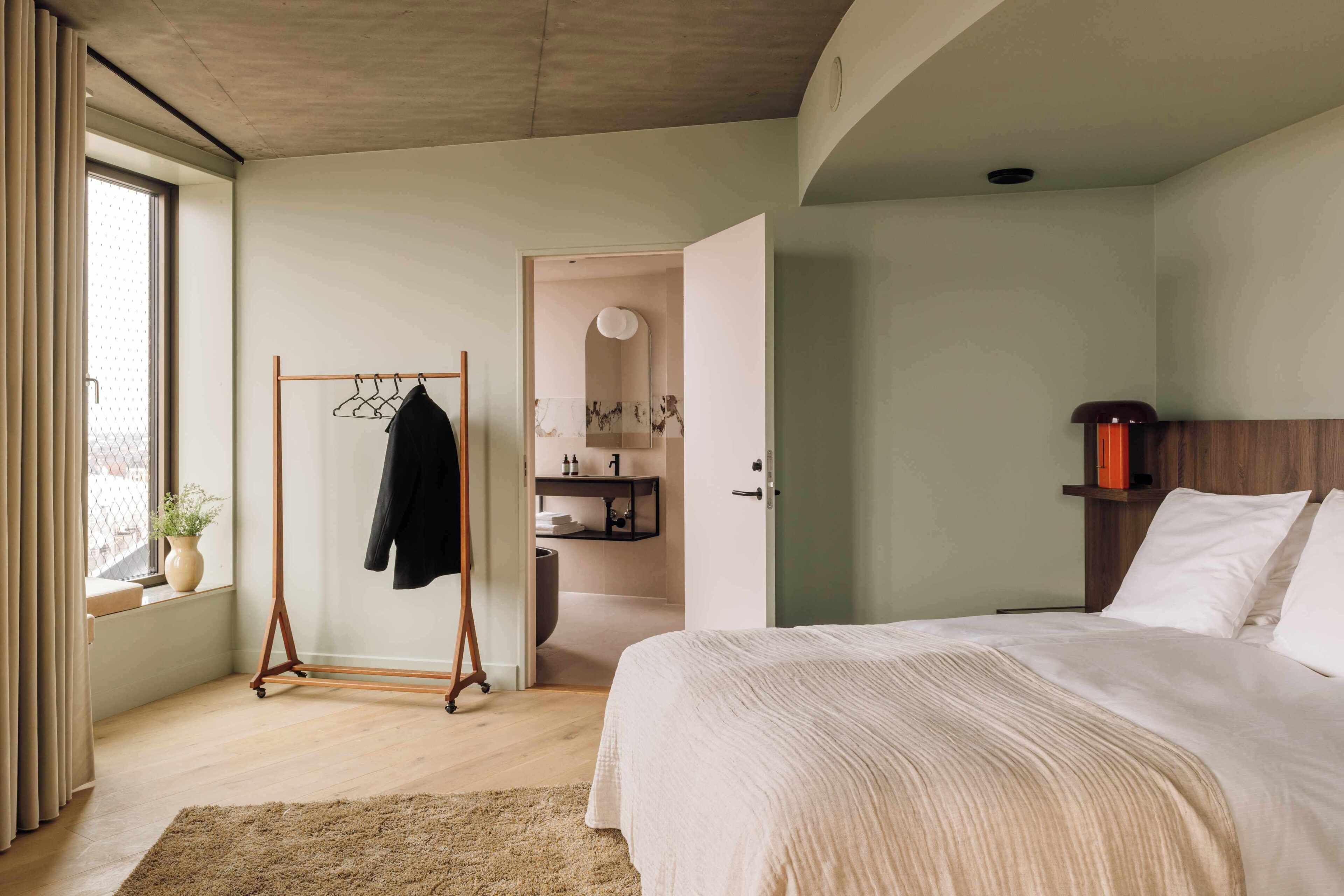 A bedroom with a bed, a wooden hanger, a bathroom with a sink and mirror, and a window with curtains.