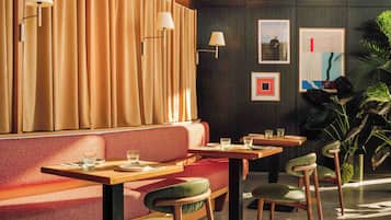 Een restaurant met een rode gestoffeerde bank, houten tafels en stoelen, wandlampen en kunstwerken in lijsten.
