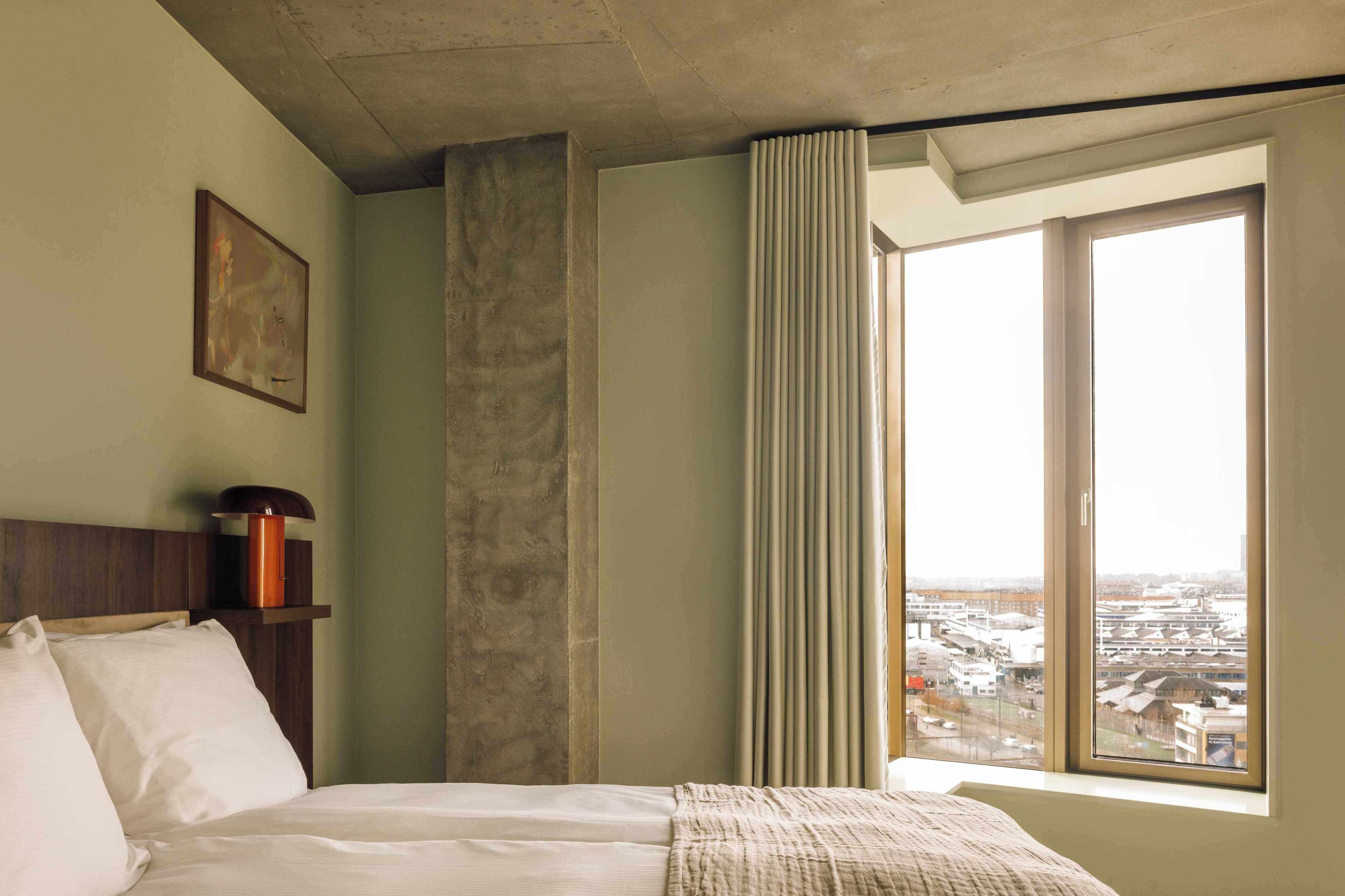 Ein Hotelzimmer mit Bett, Nachttisch, Lampe, einem Fenster mit Blick auf die Stadt und einem Vorhang.
