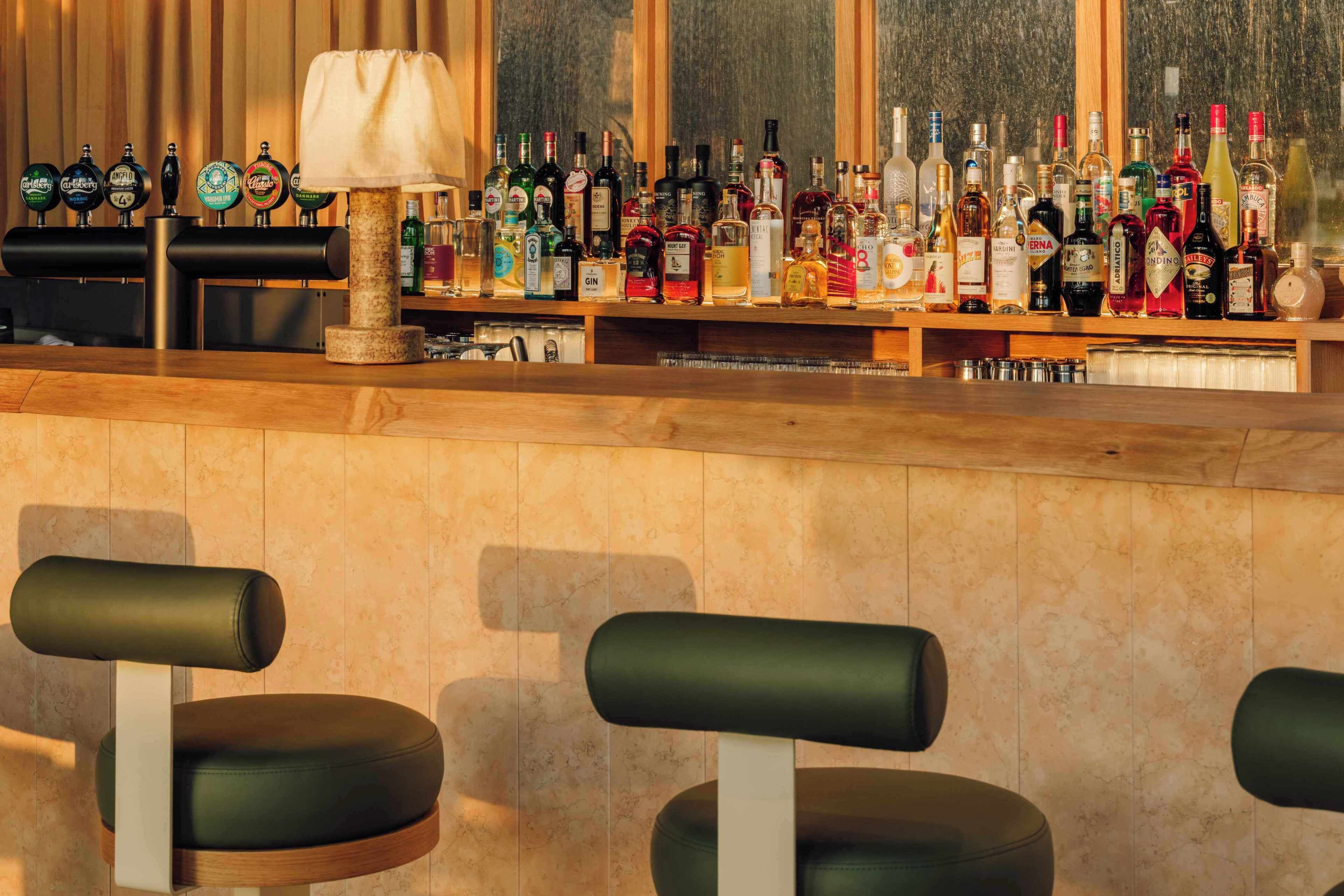 A bar area with stools, a variety of bottles, and a window with blinds.