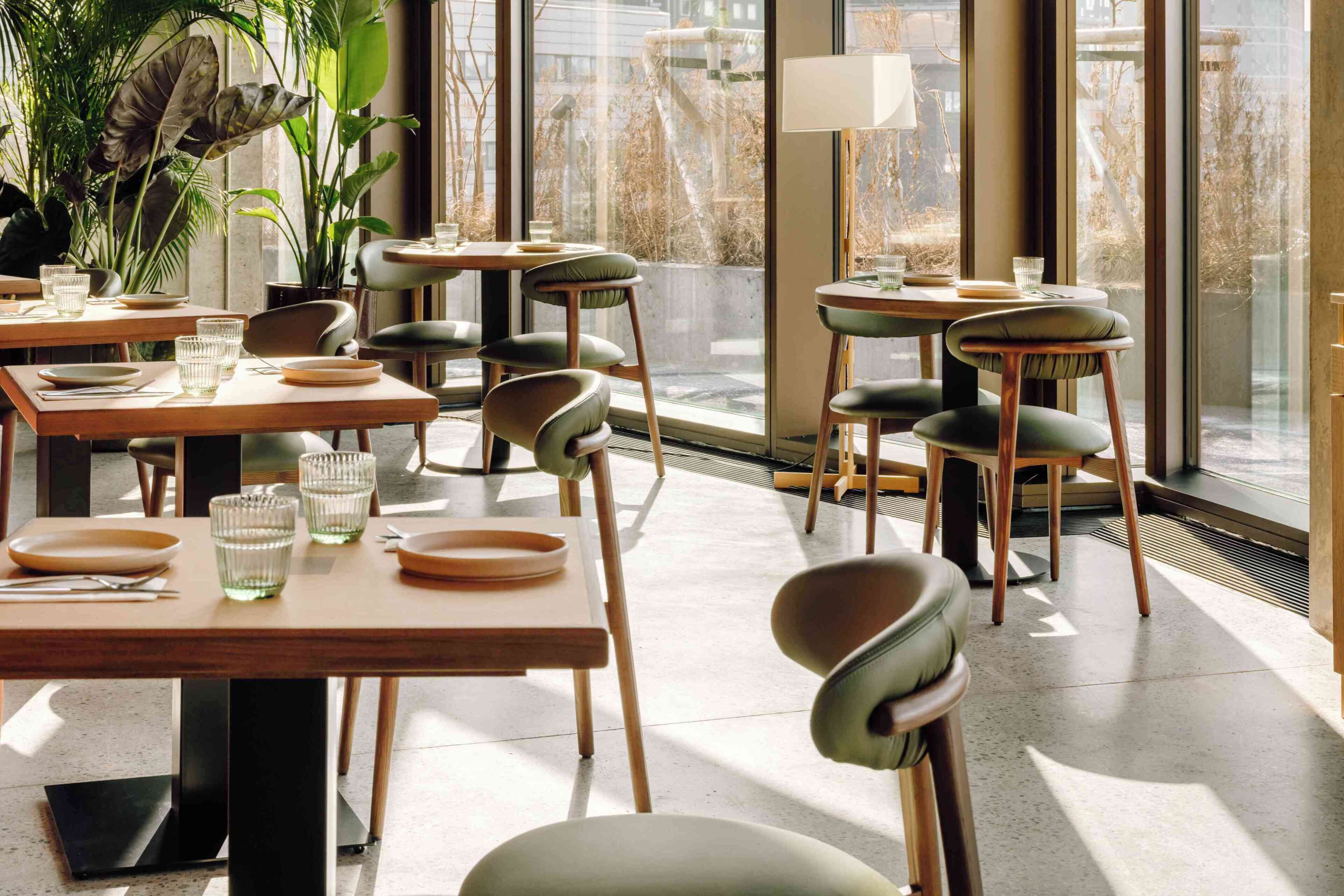 L’intérieur d’un restaurant moderne, avec des tables en bois, des chaises rembourrées de couleur verte et de grandes fenêtres offrant une vue sur la ville.