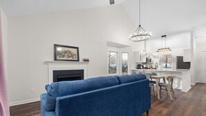 A modern living room with a blue sofa, a fireplace, and a kitchen area with white cabinets and a dining table. - NEW Fully Remodeled Home w/ Office Workspace, Patio, 3 Bed, 2 Bath (Holly Springs)