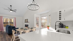 A modern kitchen with a large island, white cabinetry, and a chandelier. There is a dining area with a table and chairs, and a blue sofa in the living room. - NEW Fully Remodeled Home w/ Office Workspace, Patio, 3 Bed, 2 Bath (Holly Springs)