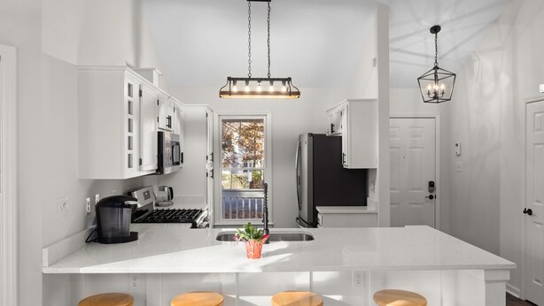 A modern kitchen with white cabinetry, a large island with stools, a hanging light fixture, and a view of the outdoors. - NEW Fully Remodeled Home w/ Office Workspace, Patio, 3 Bed, 2 Bath (Holly Springs)