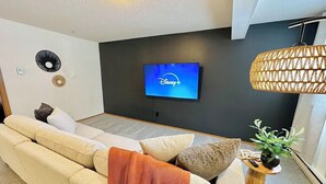Un salon moderne avec un canapé beige, un téléviseur à écran plat diffusant Disney+, un luminaire en fibres tressées et une petite table d’appoint en bois sur laquelle est posée une plante.