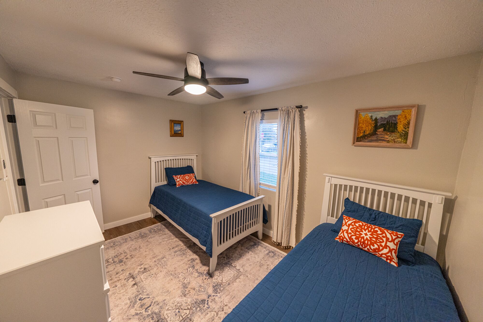 A bedroom with two beds, a ceiling fan, a window with curtains, and a painting on the wall.