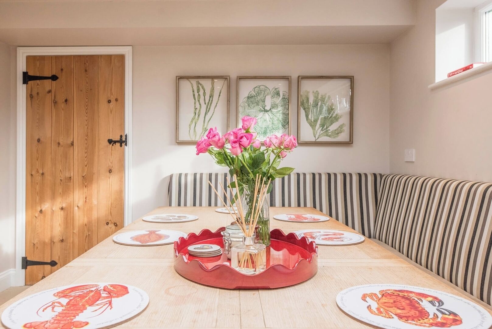 Comedor con mesa de madera dispuesta para cuatro personas, un florero con flores rosas y cojines a rayas.