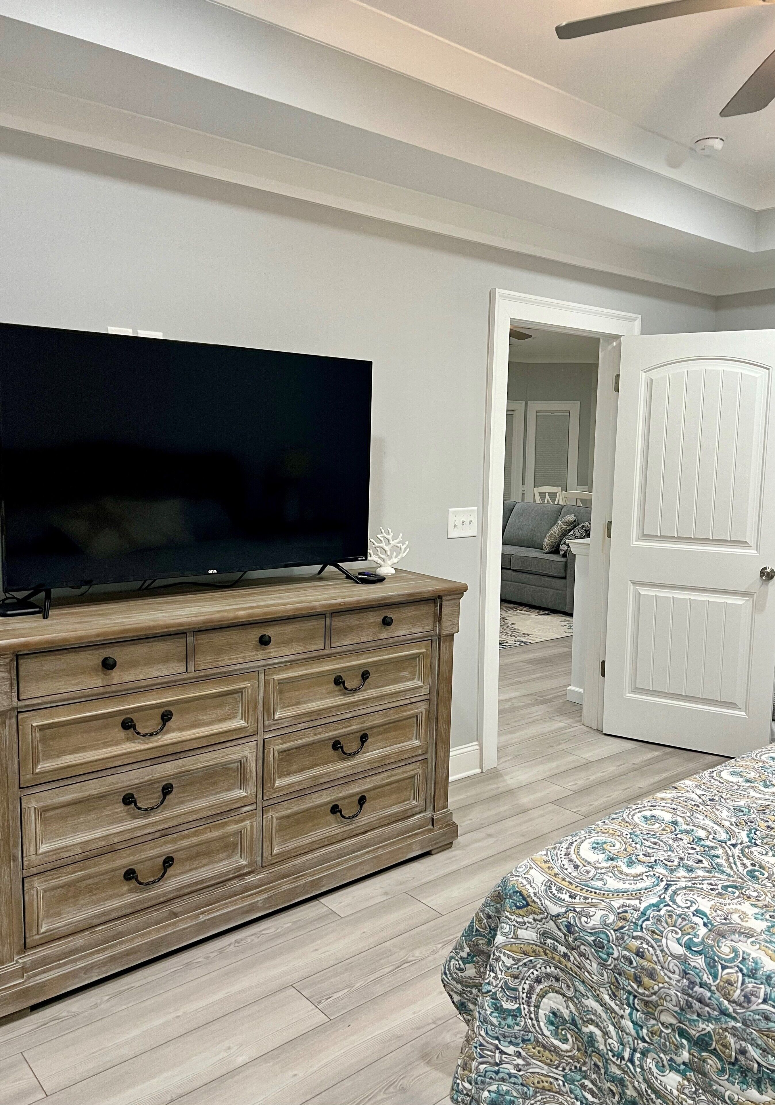 A bedroom with a large flat-screen TV on a wooden dresser, a patterned bedspread, and a hallway leading to another room.
