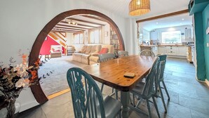 A spacious living area with a wooden dining table, teal chairs, and a large arched doorway leading to a kitchen and a cozy seating area. - The Old Barn near St Ives - Rustic Charm & Space (Saint Ives)