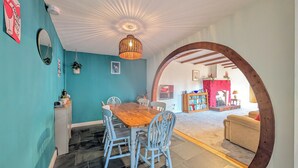 A dining area with a wooden table and blue chairs, a bookshelf, and a fireplace in the background. - The Old Barn near St Ives - Rustic Charm & Space (Saint Ives)