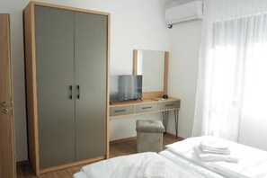Une chambre d’hôtel équipée d’un placard, d’un bureau avec une télévision, d’un lit avec du linge de maison blanc et d’une fenêtre avec des rideaux légers.