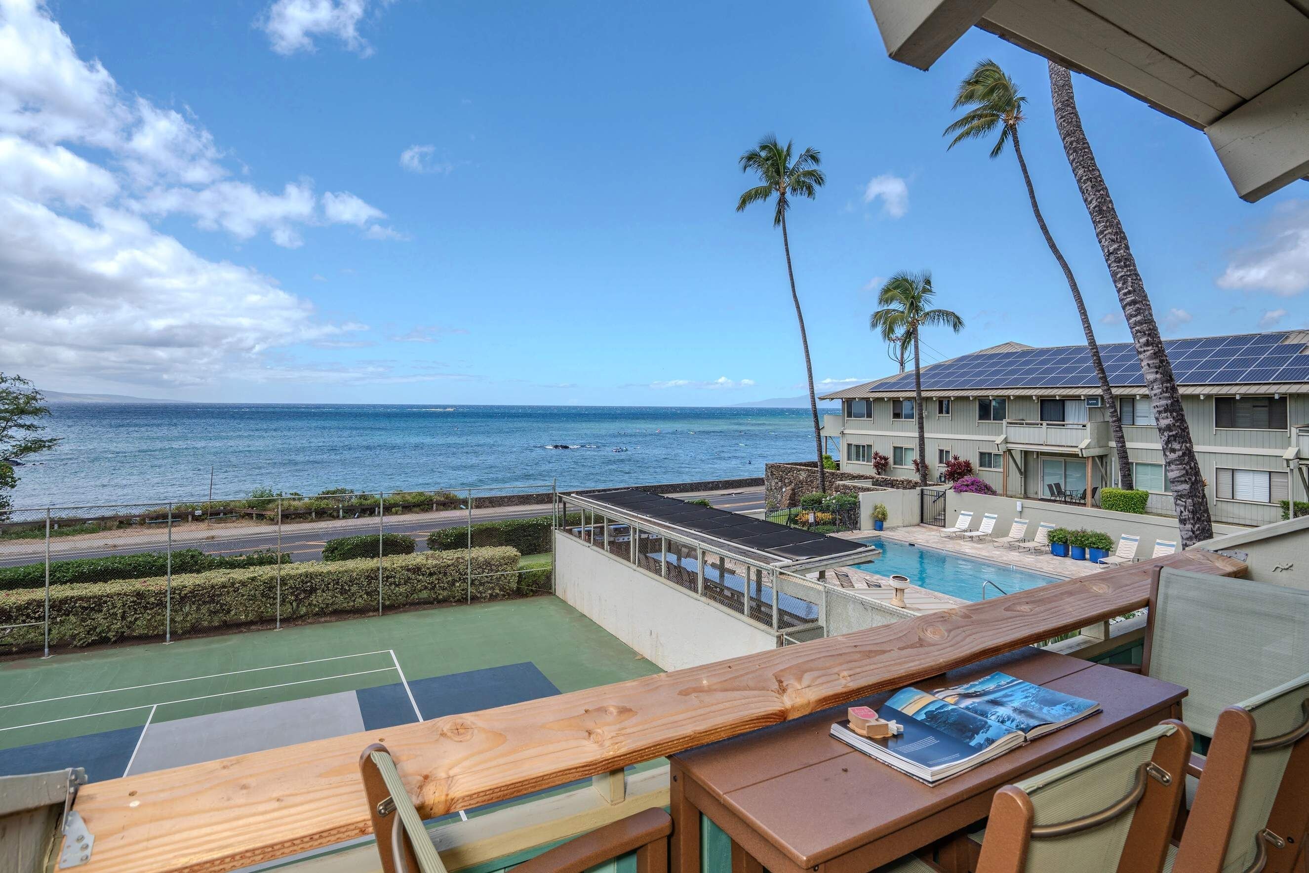 A pool area with a view of the ocean, a tennis court, and a building with solar panels.