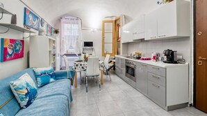A compact kitchen with white cabinets, a dining area with a table and chairs, and a blue sofa with decorative pillows. - Oltremare - Sanremo Studio Near Casino, Sanremo, Italy (Sanremo)