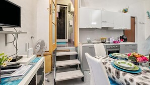 A compact kitchen with white cabinets, a dining table set with plates and glasses, a TV mounted on the wall, and a fan on the counter. - Oltremare - Sanremo Studio Near Casino, Sanremo, Italy (Sanremo)