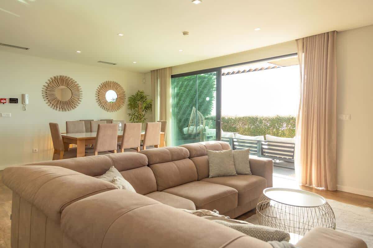A modern living room with a sectional sofa, a dining table with chairs, a potted plant, and a large sliding glass door leading to an outdoor area.