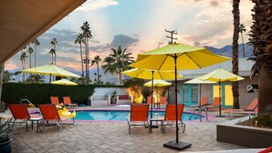 A poolside area with multiple lounge chairs and umbrellas, a swimming pool, and palm trees in the background.