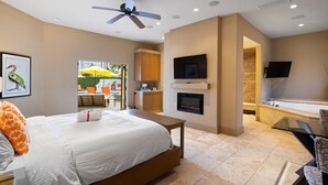 A hotel room with a bed, a ceiling fan, a fireplace, a TV mounted on the wall, and a bathroom with a bathtub and shower.