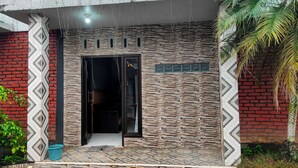 A building entrance with a patterned facade, a door with a number sign, and a small garden area with plants and a palm tree. - Hotel Intan Niaga (Banjaran)