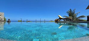 Une piscine aux eaux cristallines offrant une vue sur l’océan, un fauteuil de relaxation et une statue.