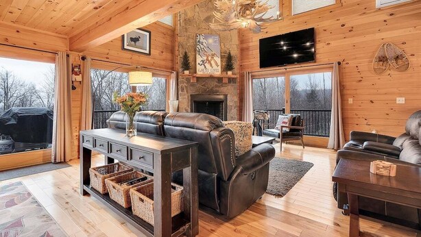 A cozy cabin interior with a fireplace, leather sofas, a TV, and a stone wall. - Mt Lodge | Sunday River Views | Hot Tub | Game Room (Newry)