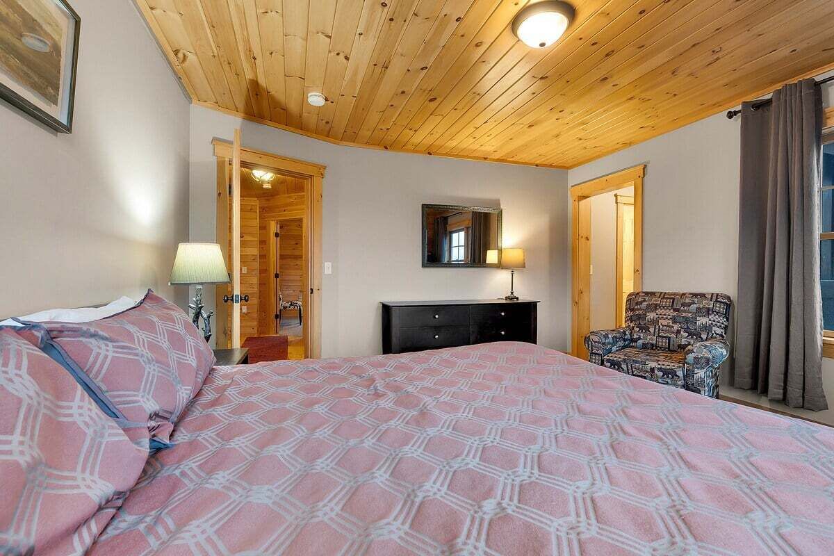 A bedroom with a wooden ceiling, a bed with patterned bedding, a dresser, a chair, and a mirror on the wall.