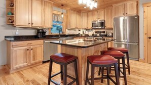 A kitchen with wooden cabinets, a stainless steel refrigerator, a coffee maker, and a large island with stools. - Mt Lodge | Sunday River Views | Hot Tub | Game Room (Newry)