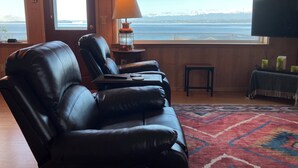 A room with a leather recliner, a lamp, a small table, a TV, and a view of the sea through large windows.