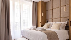 A hotel room with a large bed, a bench, a wooden headboard, and a view of the outside through sheer curtains.