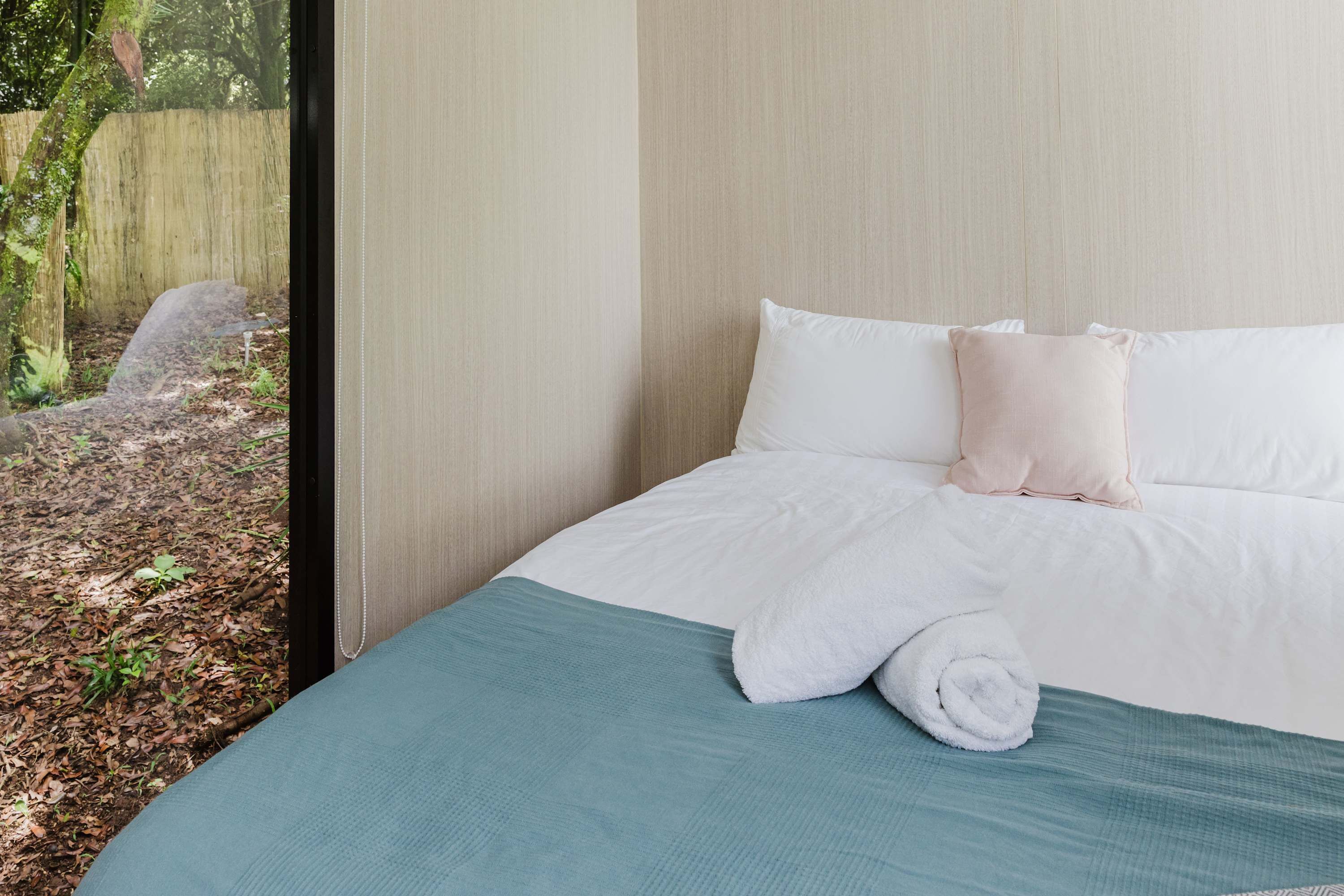Un lit bien fait, avec du linge de lit blanc et une couvre-lit turquoise, une fenêtre donnant sur une clôture en bois et de la verdure, et une tête de lit en bois.