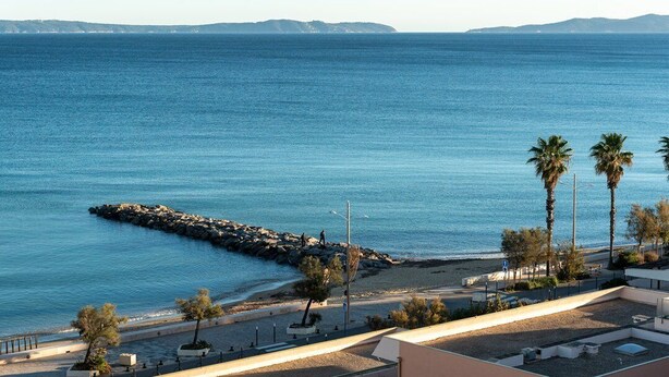 A coastal view with a rocky breakwater, palm trees, and a clear blue sea. - Location d'un studio cabine pour 4 personnes face mer, centre ville. (LE LAVANDOU)