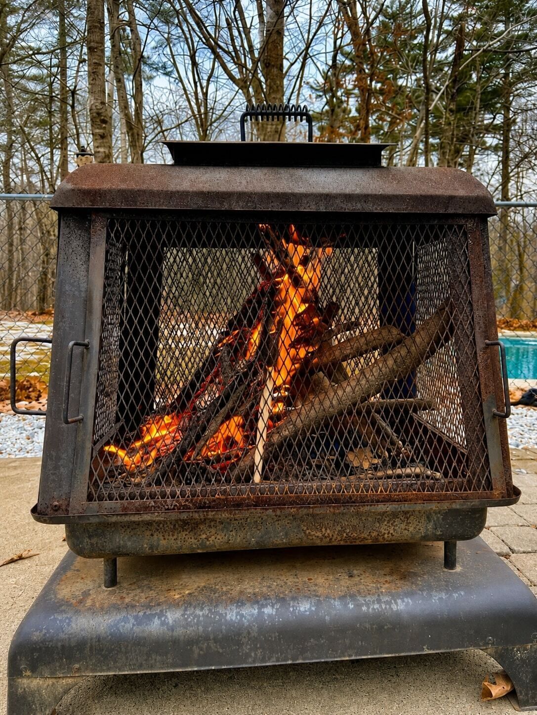 A metal fire pit with a mesh front, logs burning inside, and a metal lid on top.