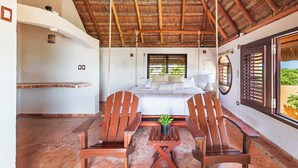 Een slaapkamer met een bed, houten stoelen, een tafeltje, een kamerplant en een raam met luiken.