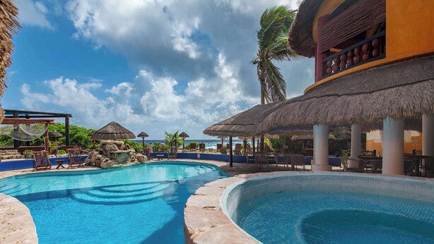 A pool area with lounge chairs and thatched umbrellas, surrounded by palm trees and a view of the ocean. - Bella Mar: Ocean Front w/ Pool Views (Isla Mujeres)