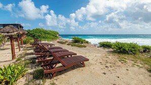 A beach with wooden lounge chairs and a thatched-roof structure. - Bella Mar: Ocean Front w/ Pool Views (Isla Mujeres)