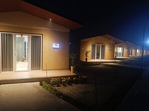 A row of modern, well-lit hotel rooms with sliding glass doors, a small outdoor area with plants, and a clear view of the sky. - Wildlense Wild Retreat (Pilibhit)