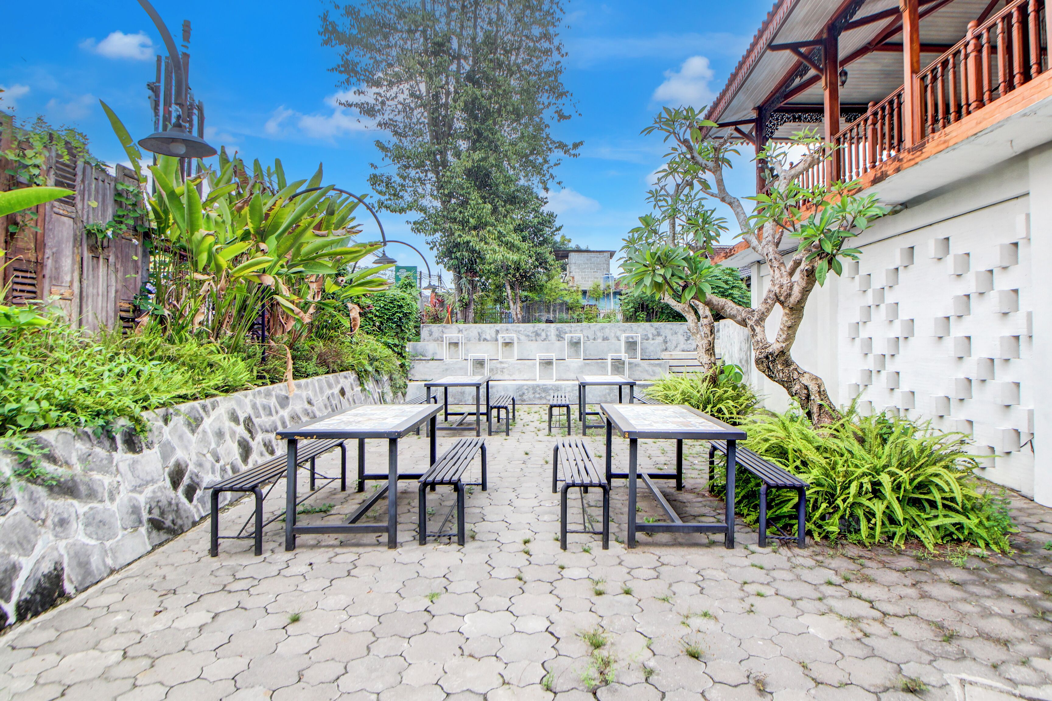 An outdoor patio with hexagonal stone tiles, black metal tables and benches, and lush greenery.