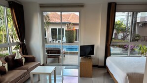 A hotel room with a sofa, a bed, a TV on a wooden stand, and a view of a swimming pool and outdoor area. - khun mai villa (Pattaya)
