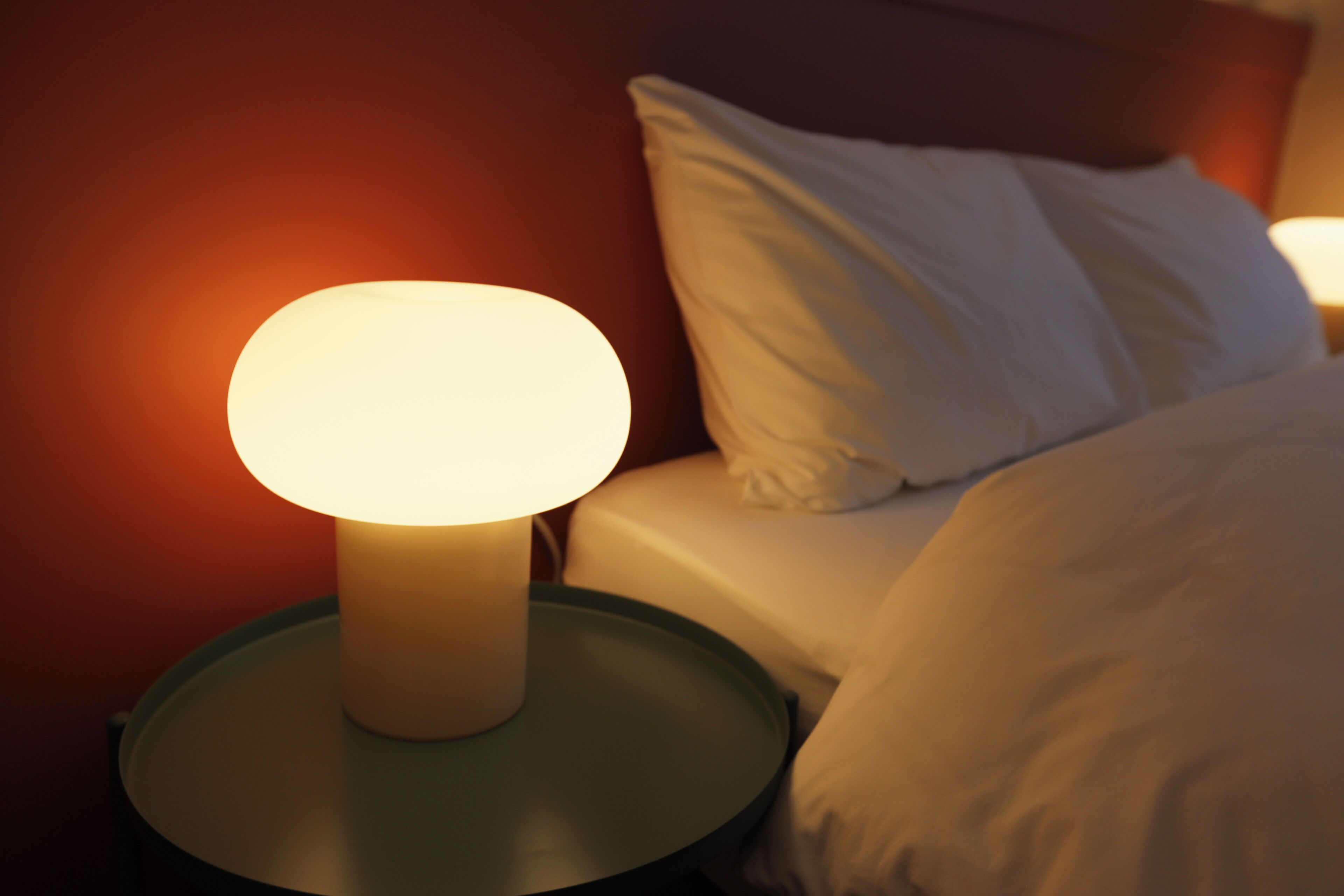 Una cama con ropa de cama y almohadas blancas, una mesita de noche con lámpara y una pared de tono cálido.