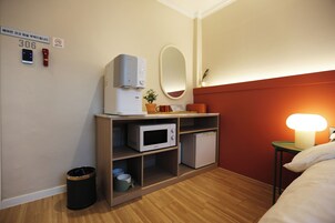 Habitación de hotel con piso de madera, cama con ropa blanca, mesita de noche con lámpara, microondas, un refrigerador pequeño, un cesto de basura y repisa con una planta y vasos.