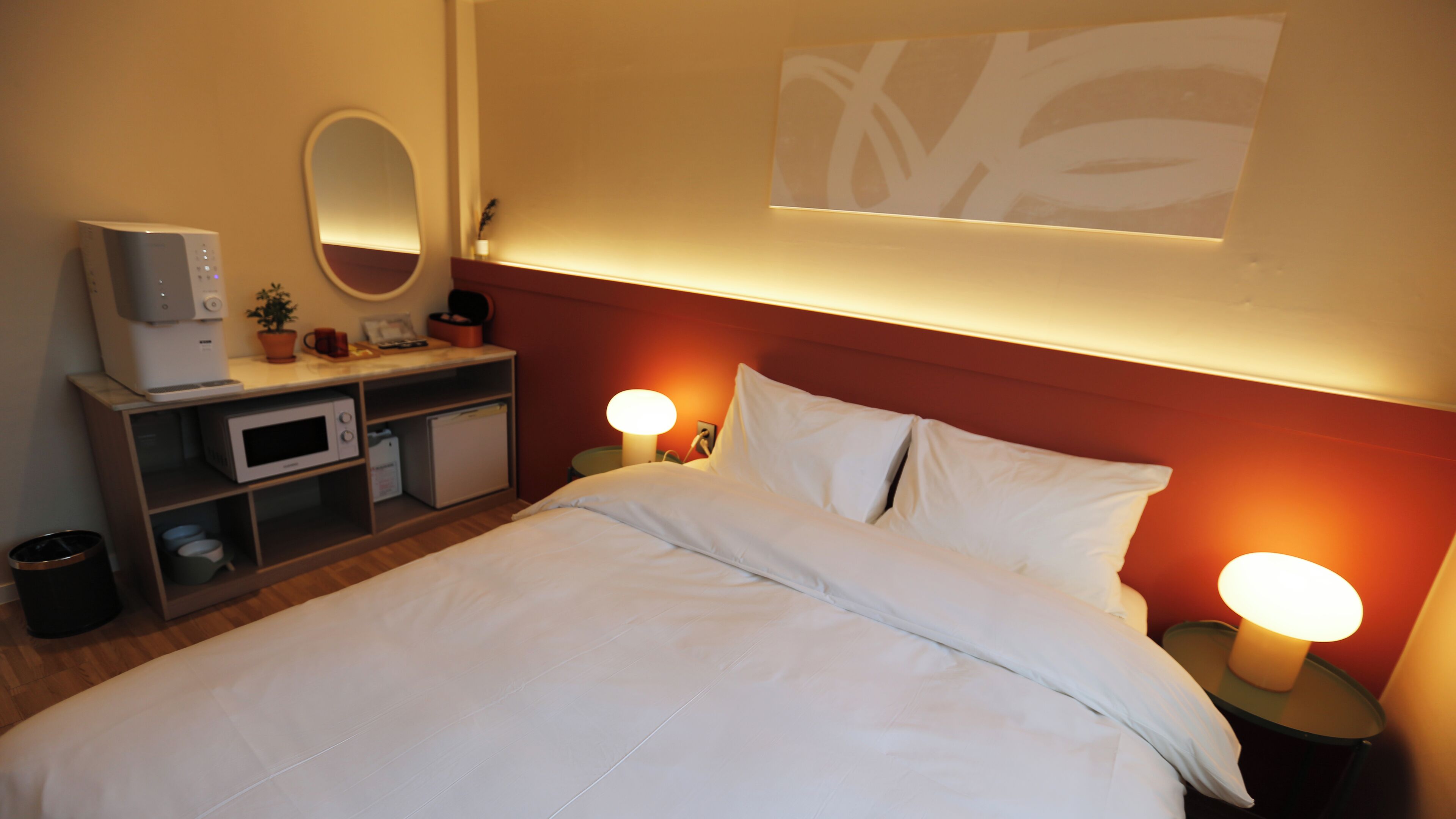 Habitación de hotel con cama, lámparas de noche, microondas, refrigerador, una mesita con espejo y una obra de arte colgada en la pared.