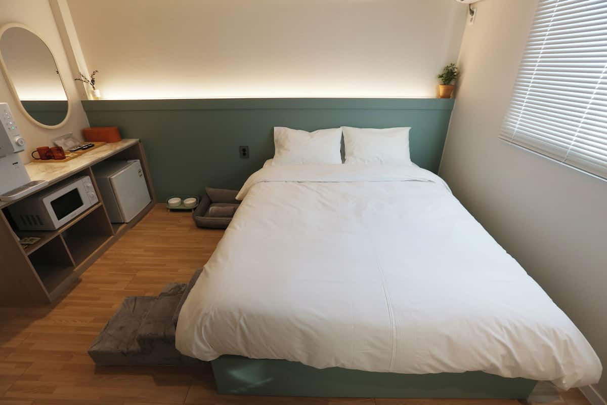 Un dormitorio ordenado con cama, mesita de noche y un pequeño espacio de escritorio con microondas y una planta.