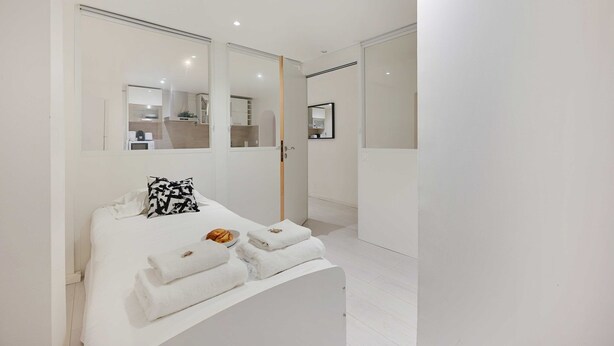 A modern bedroom with a bed, a bedside table with towels, a mirror, and a view of a bathroom and kitchen area.