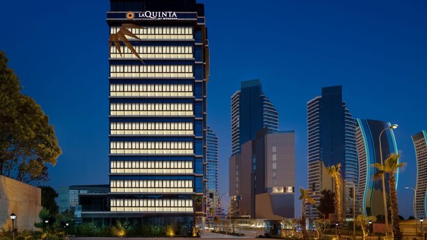 Korkea hotellirakennus, jonka julkisivussa on suuri ja näkyvästi sijoitettu kyltti, jossa lukee »La Quinta by Wyndham». Hotellin ympärillä on moderneja rakennuksia ja palmuja.