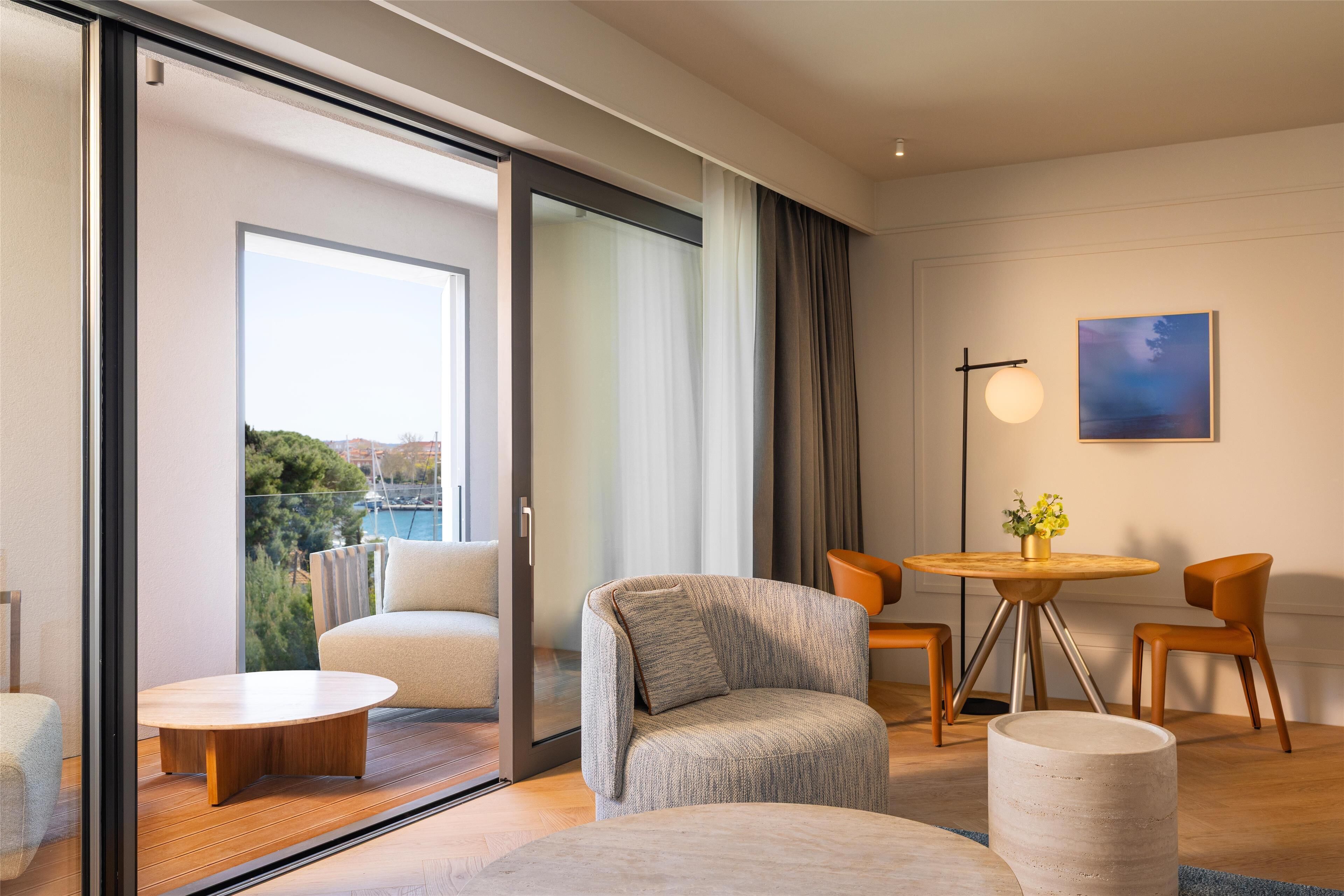 A modern living room with a sofa, chairs, a coffee table, and a dining table with chairs. There is a large window with a view of trees and buildings.