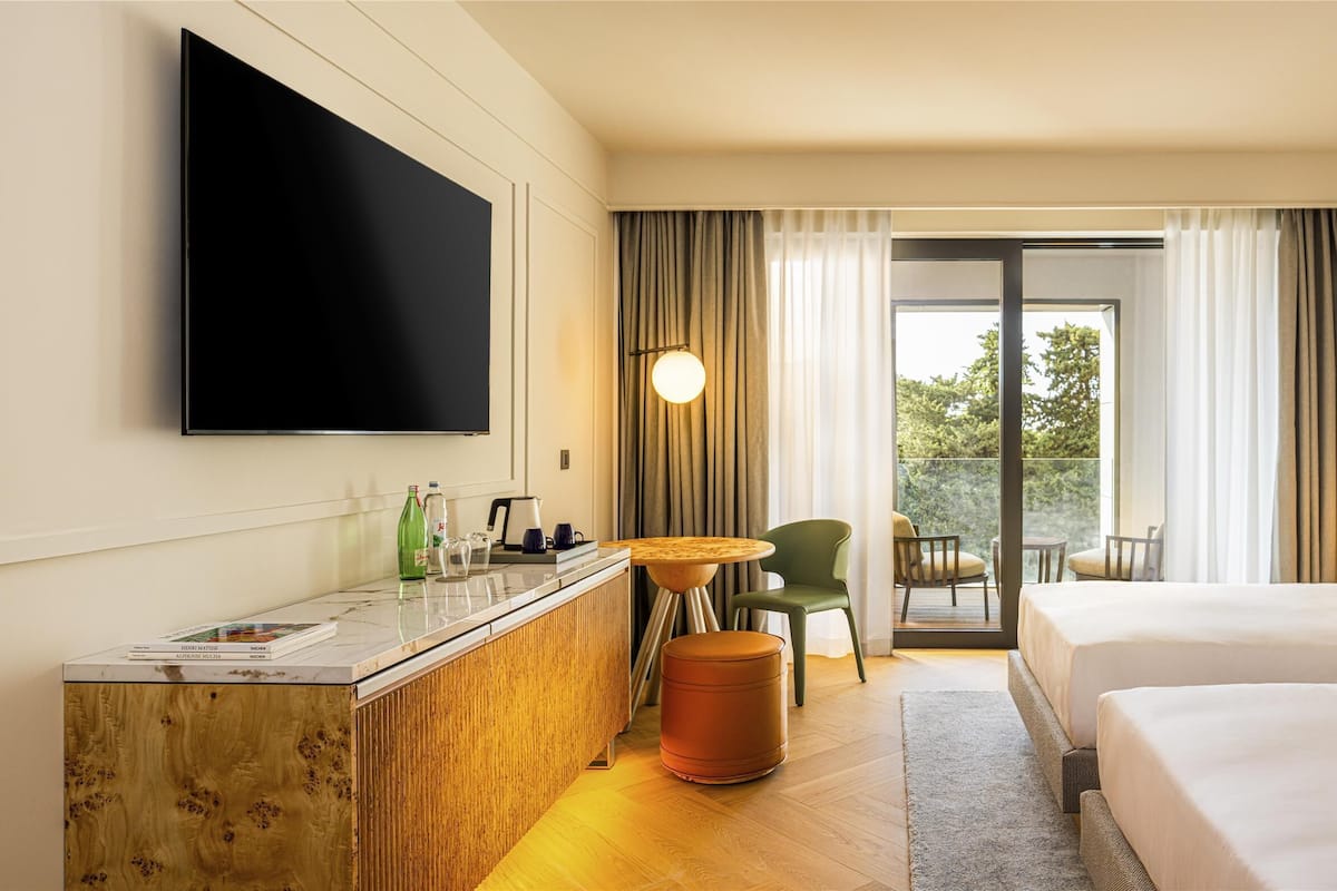 A hotel room with two beds, a desk with a flat-screen TV, a small table, and a view of greenery through the window.