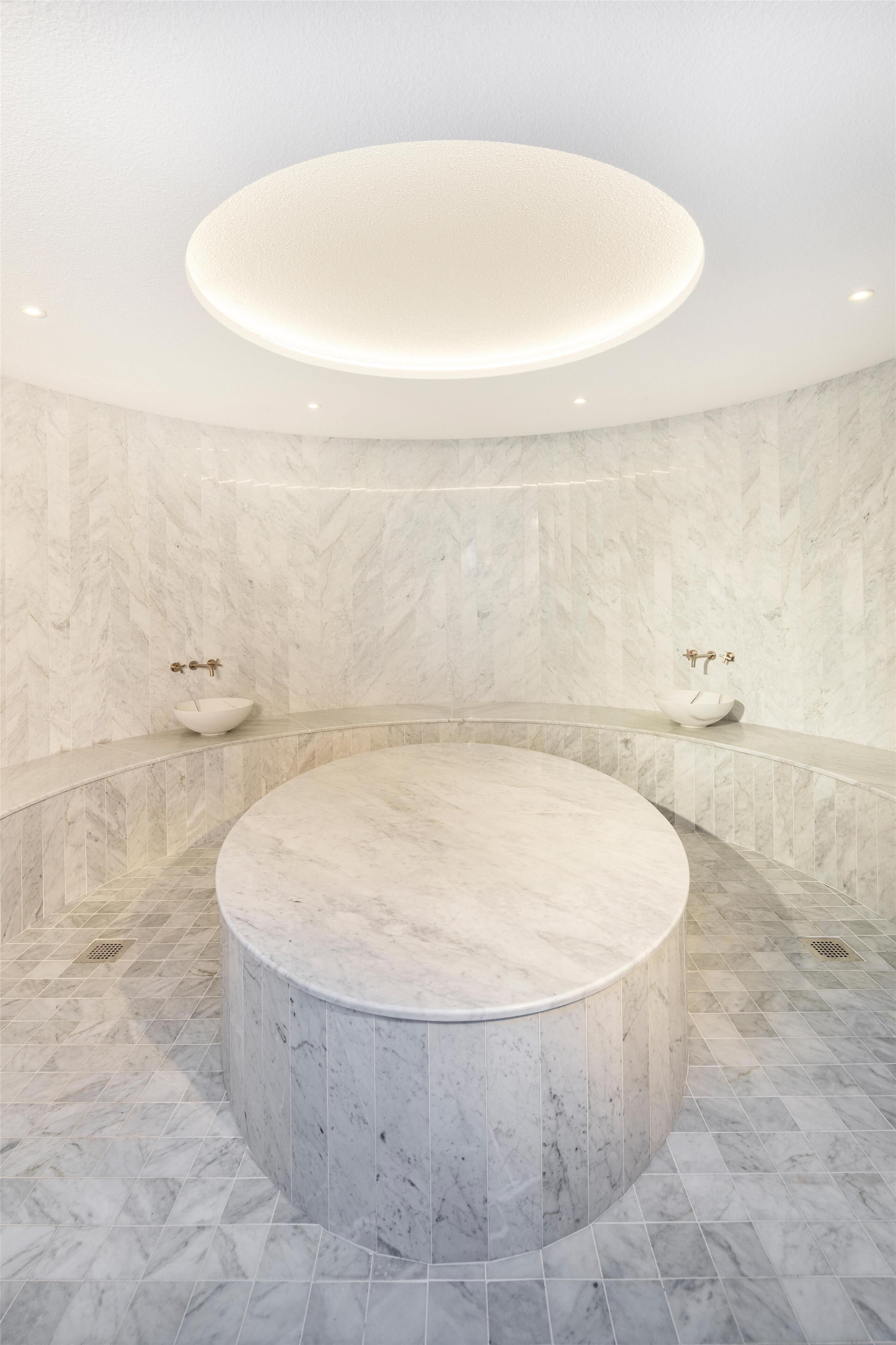 A circular marble bathtub with a central light fixture, two sinks, and marble walls.