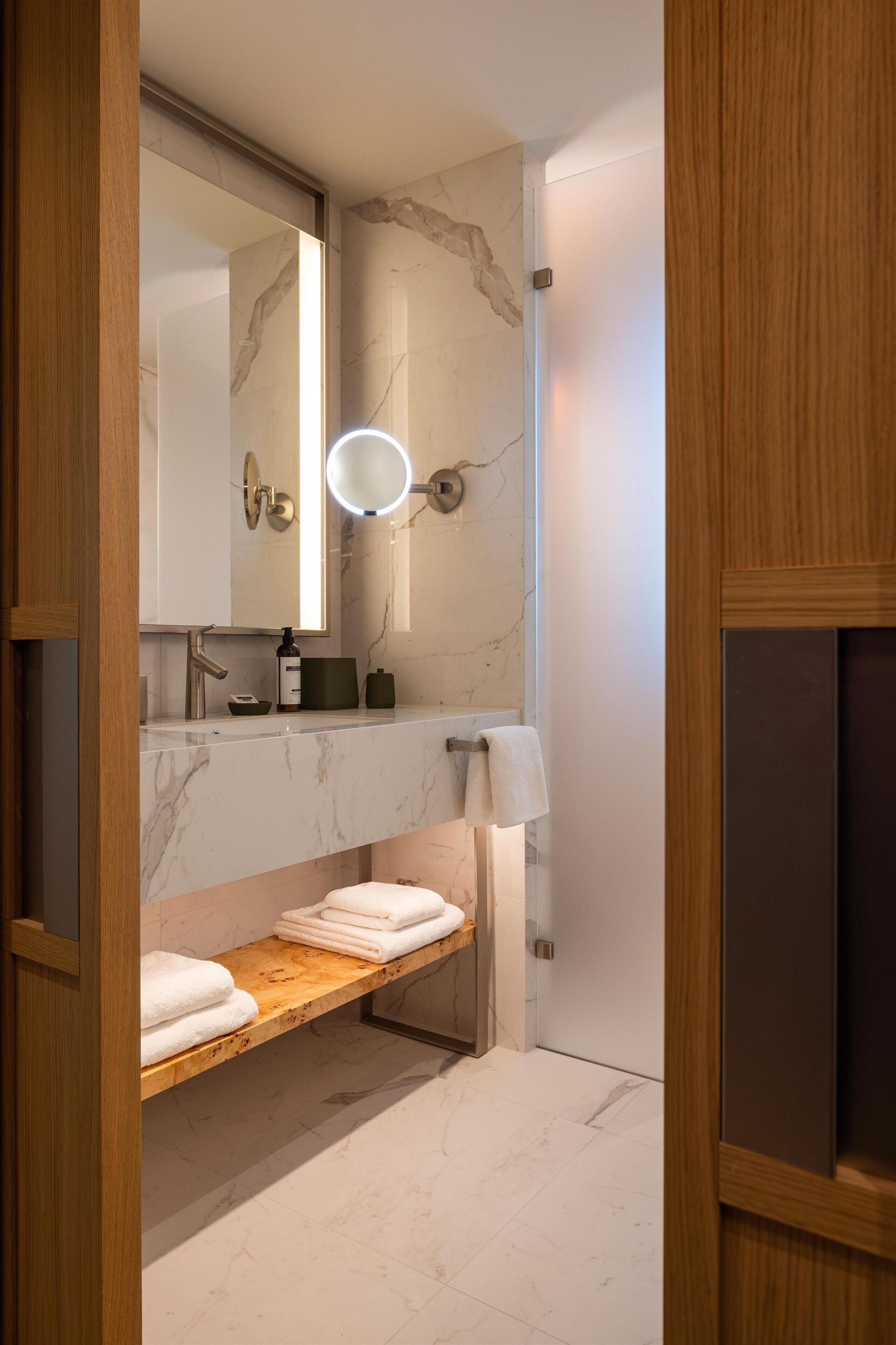 A modern bathroom with a marble vanity, a wooden shelf with towels, a round mirror with a light, and a towel rack.