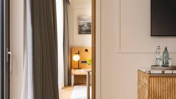 Ein modernes Hotelzimmer mit Holzboden, einem großen Fenster mit Vorhängen, einer Couch, einem kleinen Tisch und einer Kommode mit Flaschen.