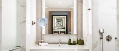 A modern bathroom with a marble vanity, a large mirror, a shower area, and a wooden shelf with towels.