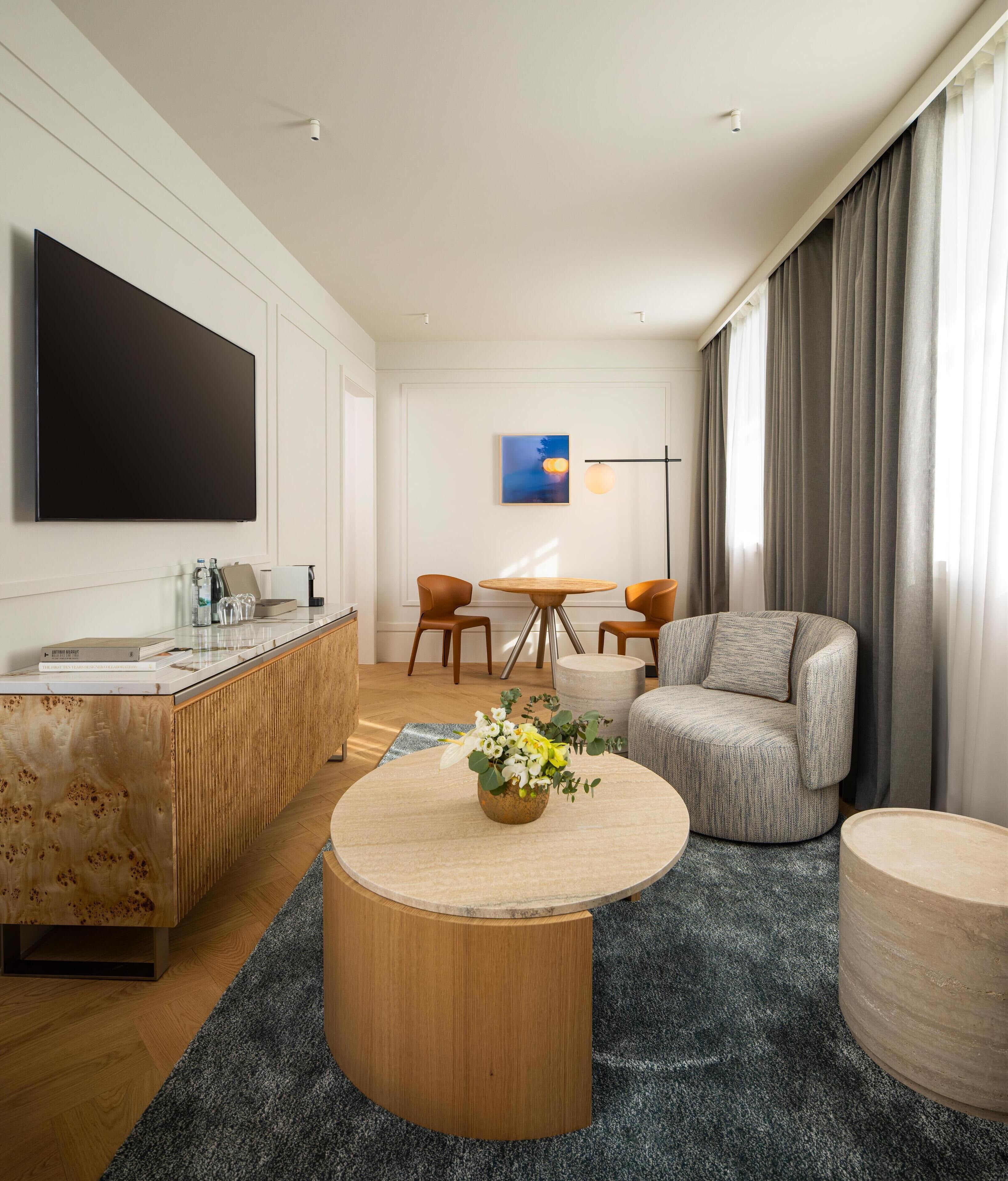 Un salon moderne avec un téléviseur à écran plat, une table basse en bois agrémentée d’un arrangement floral, un canapé gris et un coin repas avec une table ronde et des chaises.